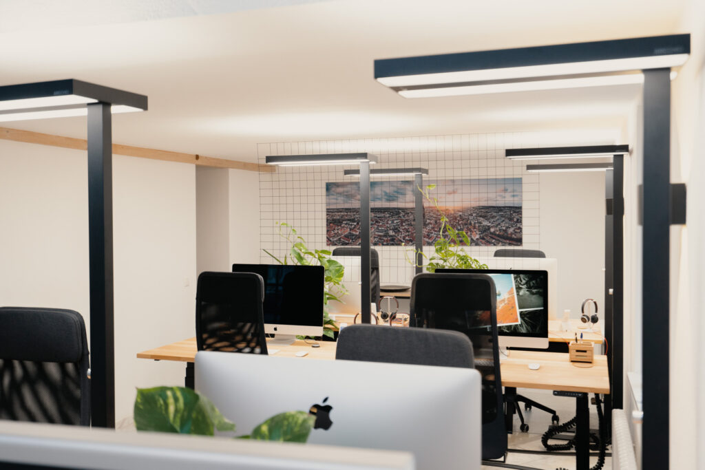 Büro mit mehreren Arbeitsplätzen, Schreibtischen, Computern und schwarzen Bürostühlen, Pflanzen und Deckenleuchten