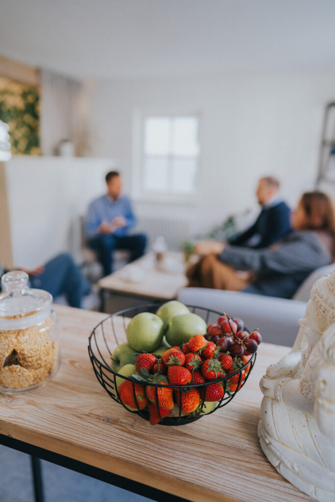Schale mit grünen Äpfeln, Erdbeeren und roten Trauben auf einem Holztisch, im Hintergrund mehrere Personen im Gespräch in einem Wohnzimmer.